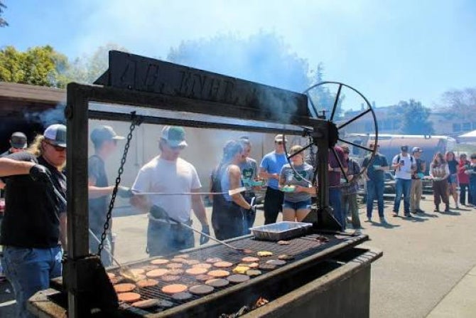 Students stand by a large barbecue pit and cook hamburgers. 