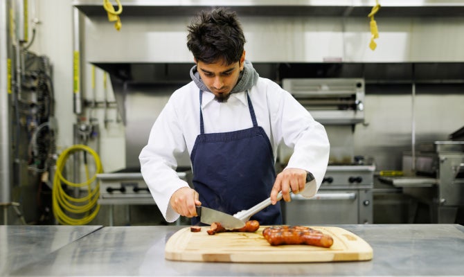 A student in a chef's coat and apron cuts into student-made sausage. 